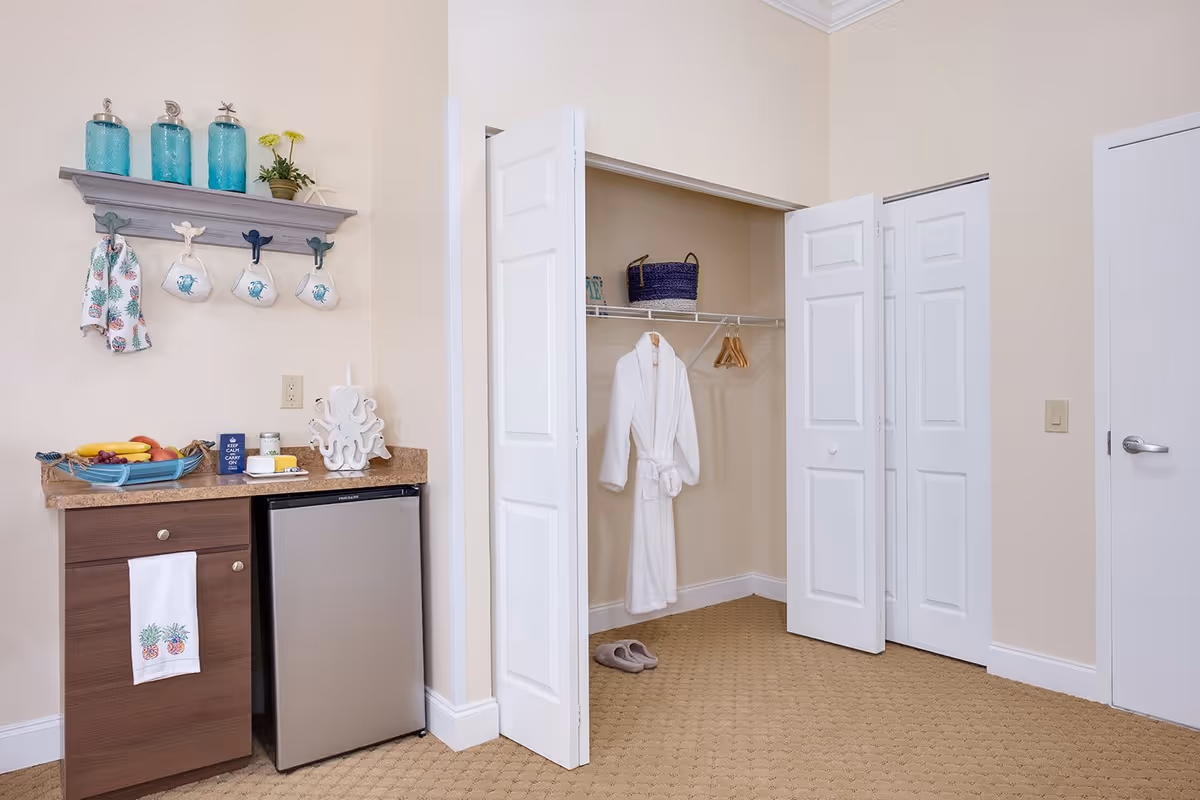 Interior of a small suite showing a countertop with a mini fridge and decorative items on the left and an open closet with a hanging robe and slippers on the right.