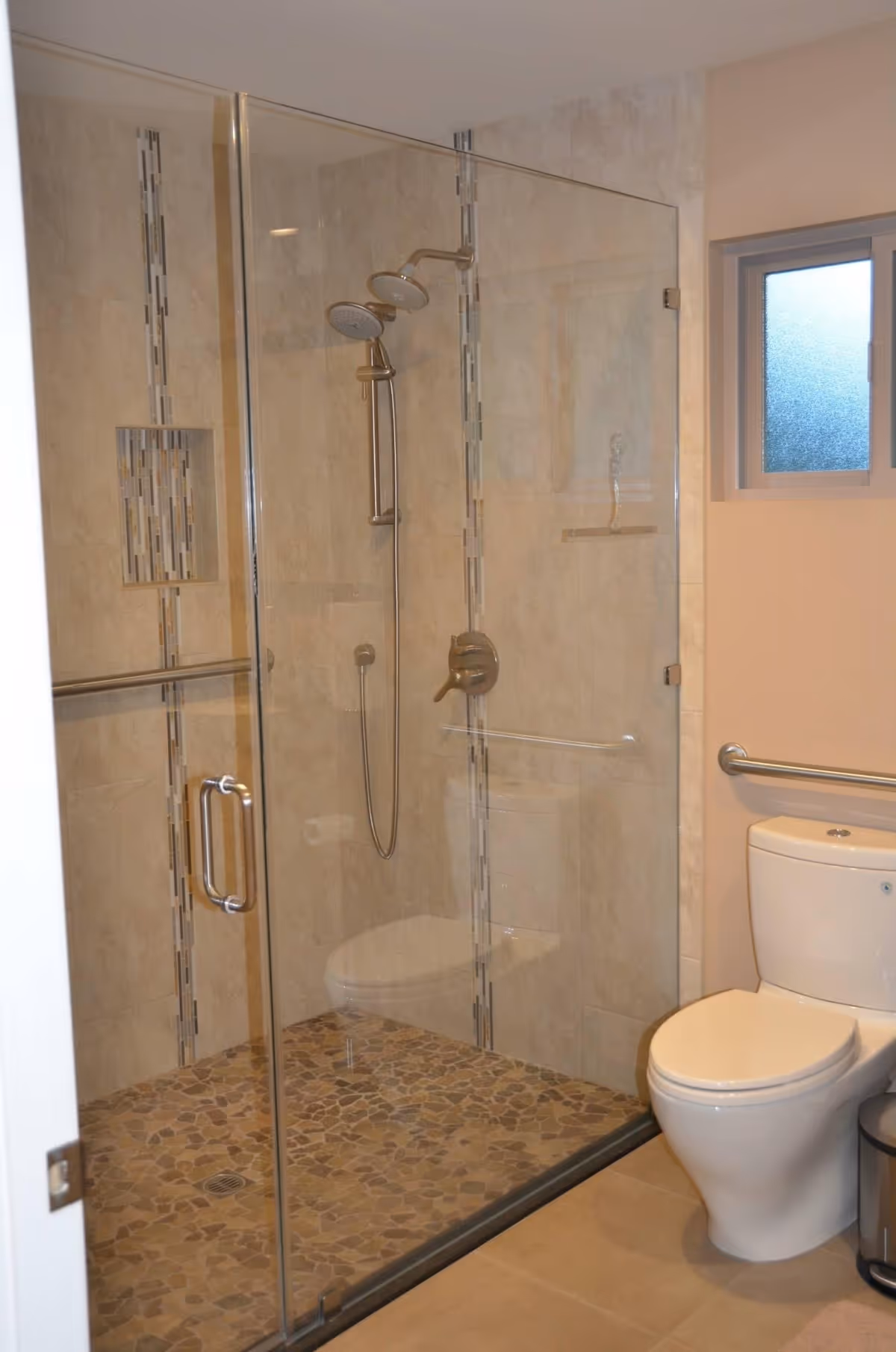 A modern bathroom featuring a glass-enclosed shower with dual showerheads and tiled walls with decorative vertical strips. The shower floor has pebble-style tiles. Next to the shower is a white toilet with a grab bar mounted on the wall beside it. A frosted window is visible above the toilet.