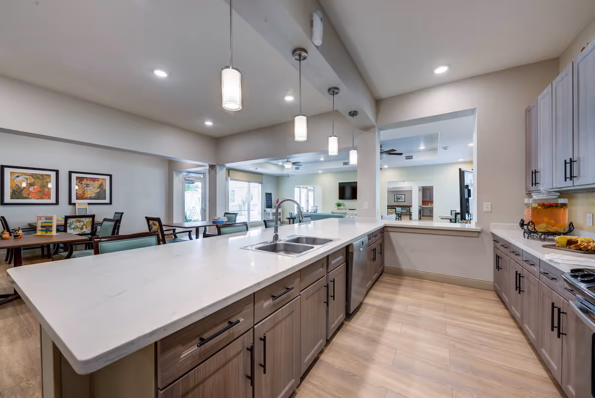 Modern kitchen area with a large white marble island countertop featuring a double sink and pendant lights hanging above. The kitchen has light wood cabinets with black handles, a dishwasher, and a beverage dispenser with fruit-infused water on the counter. In the background, there is a dining area with tables and chairs, wall art, and large windows letting in natural light.