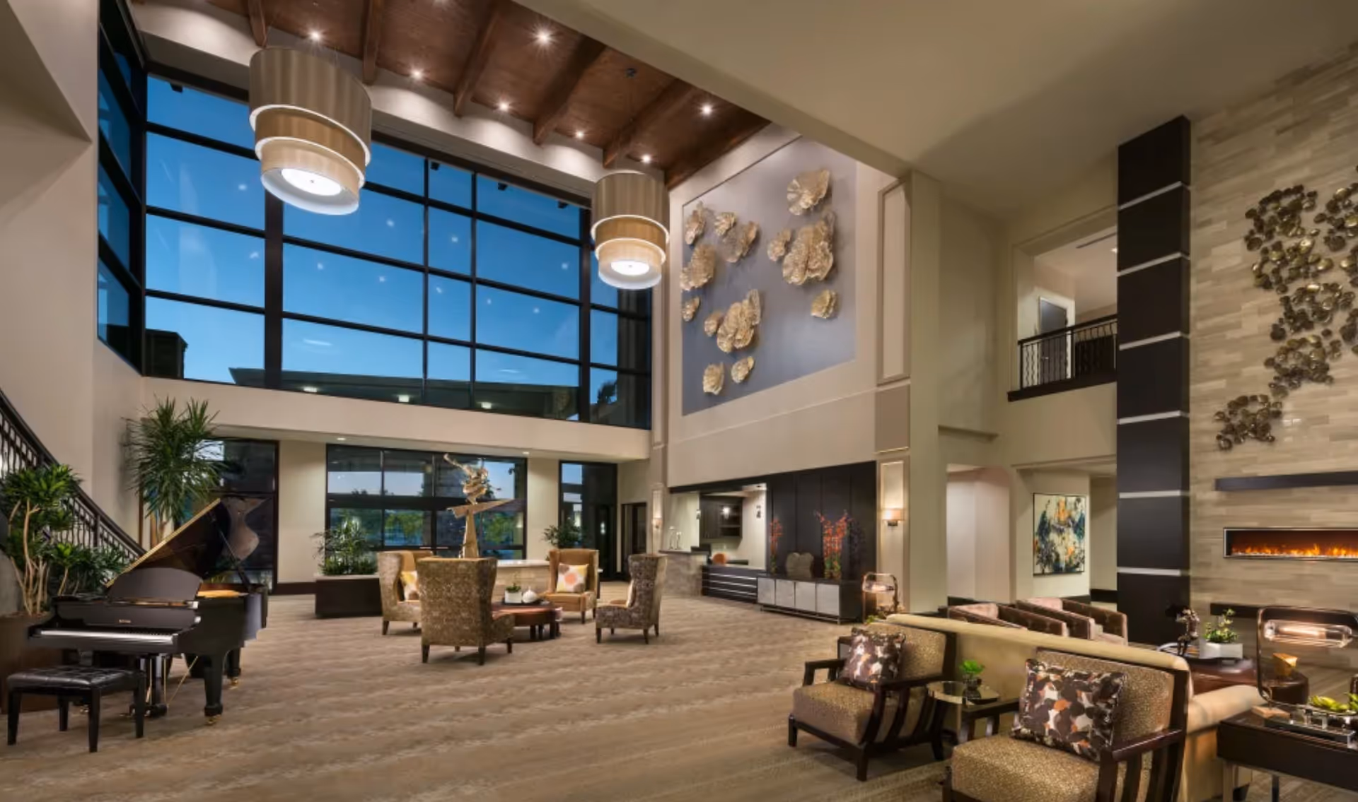 Spacious and elegant living room area with high ceilings, large floor-to-ceiling windows showing a twilight sky, modern hanging light fixtures, a grand piano, multiple seating arrangements with armchairs and sofas, decorative wall art, and a fireplace.