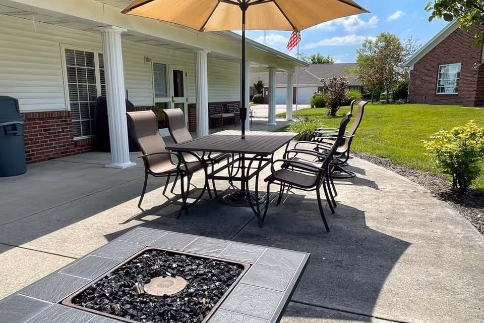 Patio with a dining table, umbrella and several chairs on a concrete terrace next to a covered walkway and lawn.