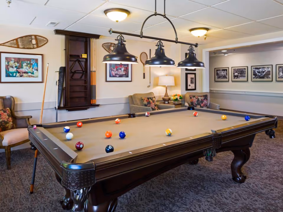 A cozy game room featuring a large pool table with billiard balls arranged on the felt surface. The room has comfortable armchairs with floral cushions, framed artwork on the walls, vintage snowshoes hanging as decoration, and a rack holding pool cues. Overhead, three black pendant lights illuminate the pool table. The carpeted floor and warm lighting create a welcoming atmosphere.
