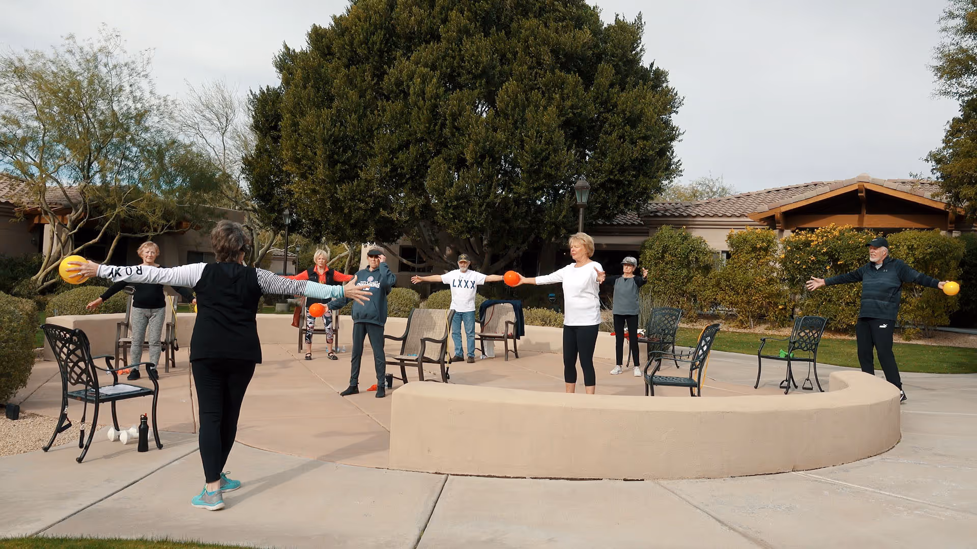 A group of senior adults participating in an outdoor exercise class at a senior living facility. They are standing in a semicircle on a paved area with chairs spaced around them, holding small exercise balls and stretching their arms out. The background shows trees, shrubs, and a building with a tiled roof.