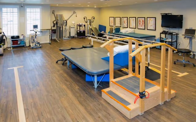 A spacious physical therapy room with exercise equipment, treatment tables, and a set of wooden therapy stairs.