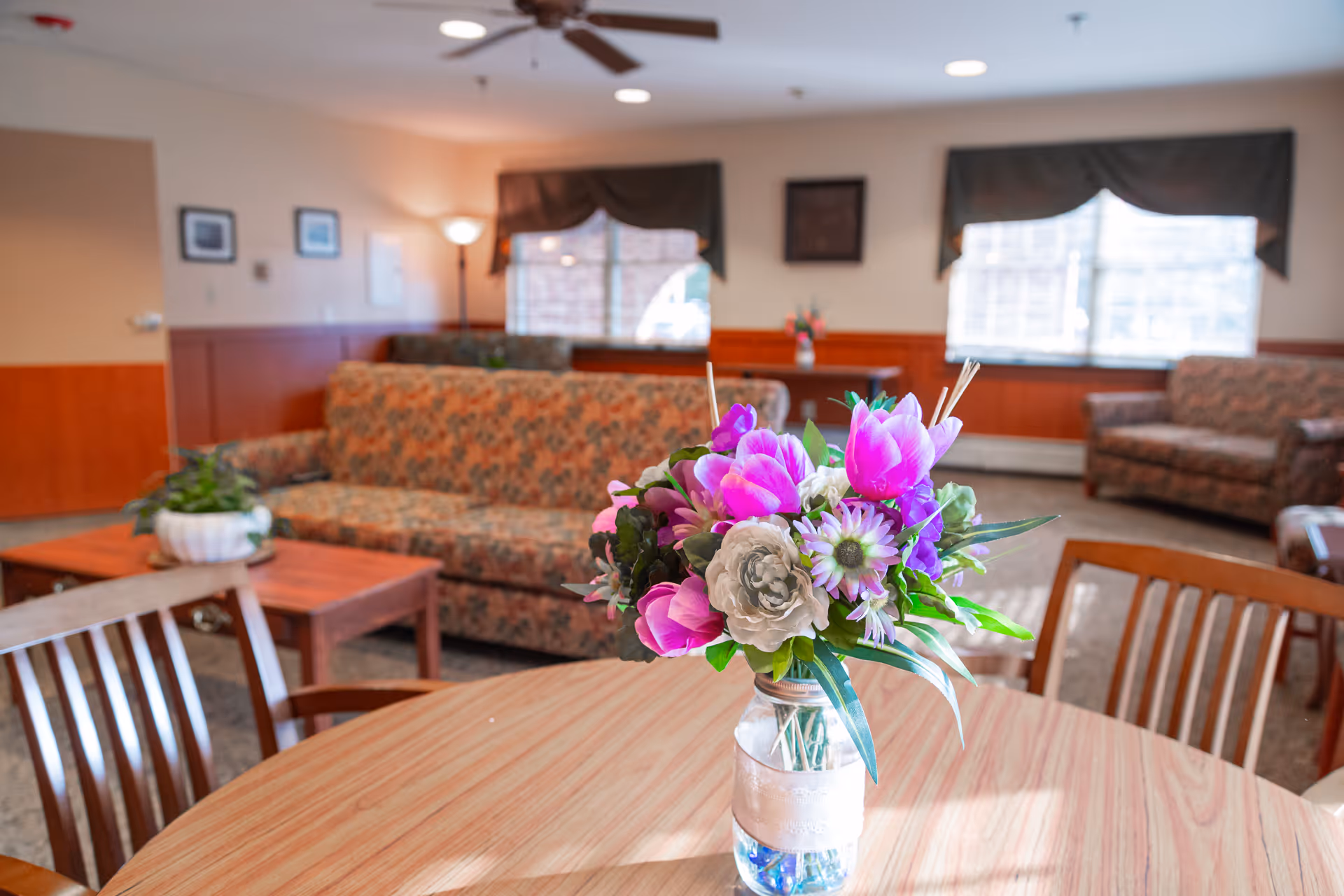 A cozy living room area in a senior care facility with floral-patterned sofas, wooden chairs around a round table with a vase of colorful flowers, large windows with dark valances, and warm lighting.