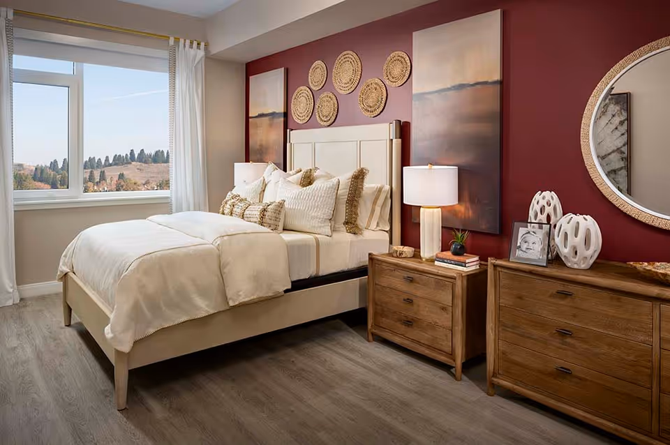 A cozy bedroom with a large bed dressed in cream-colored bedding and multiple pillows. The room features a wooden nightstand with a lamp and decorative items, a wooden dresser with a round mirror above it, and wall art including circular woven pieces and two large paintings on a deep red accent wall. A window with white curtains offers a view of trees and hills outside.