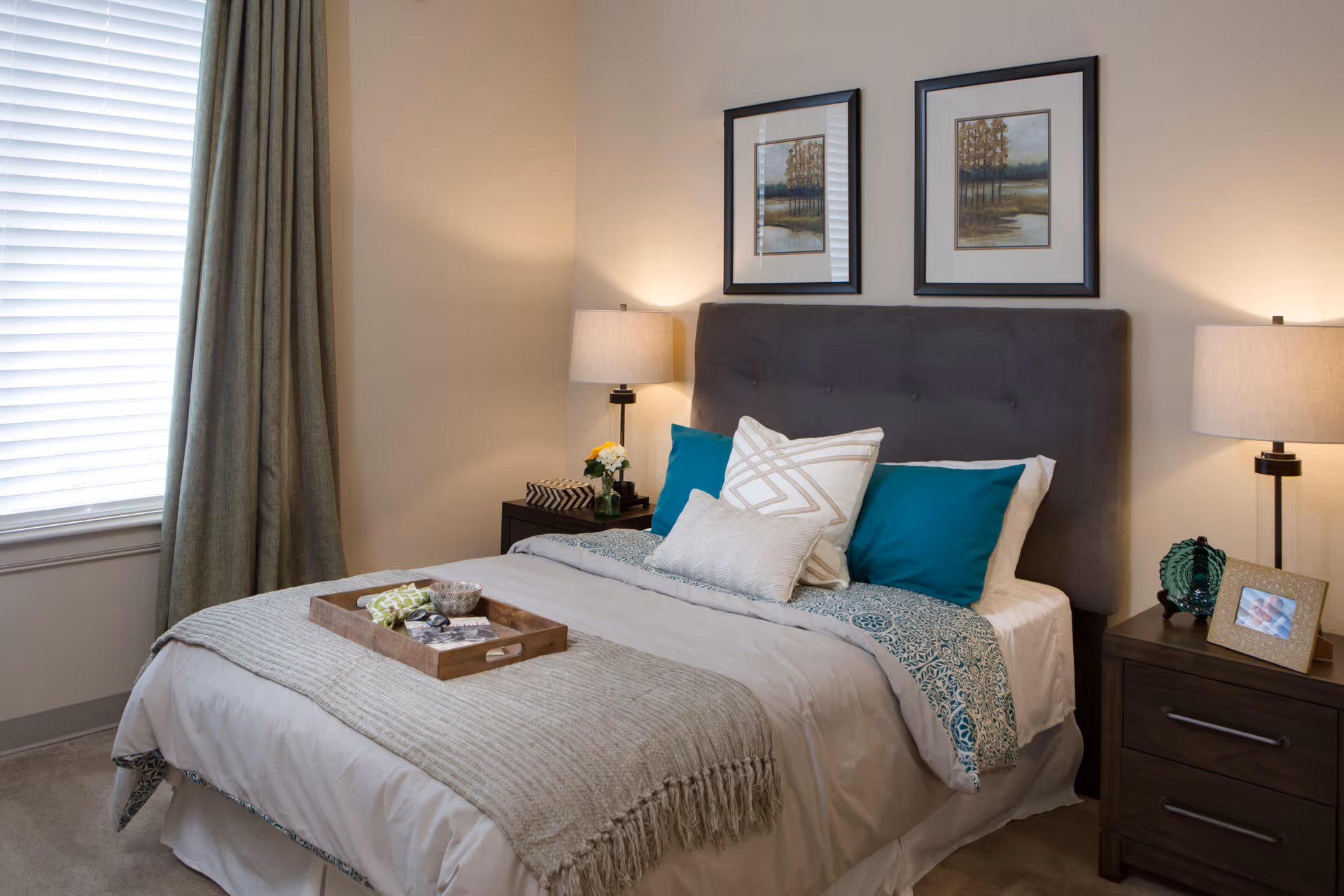 A neatly made bed with a gray upholstered headboard, white and teal pillows, and a beige throw blanket. There is a wooden tray with decorative items on the bed. On either side of the bed are dark wood nightstands with lamps, a small plant, and a framed photo. Two framed landscape paintings hang above the headboard. A window with closed blinds and green curtains is visible on the left side.