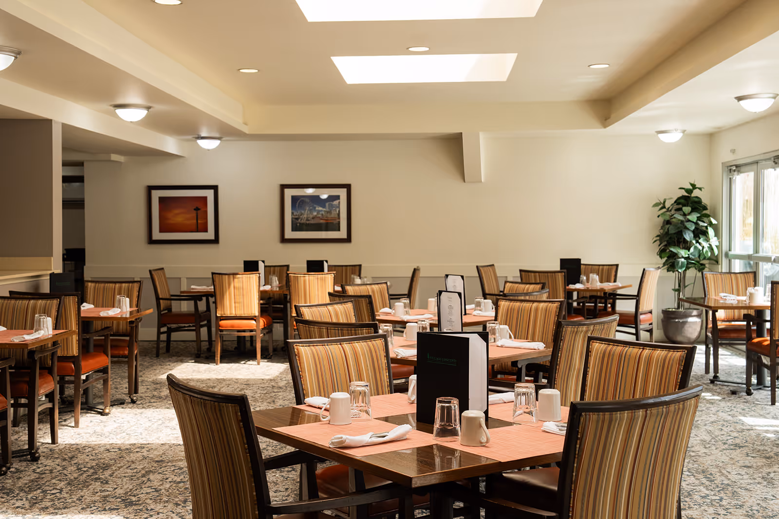 A well-lit dining room with multiple square wooden tables set with placemats, napkins, cups, and glasses. The chairs have striped upholstery with orange cushions. There are framed pictures on the wall and a large potted plant near the glass doors letting in natural light.