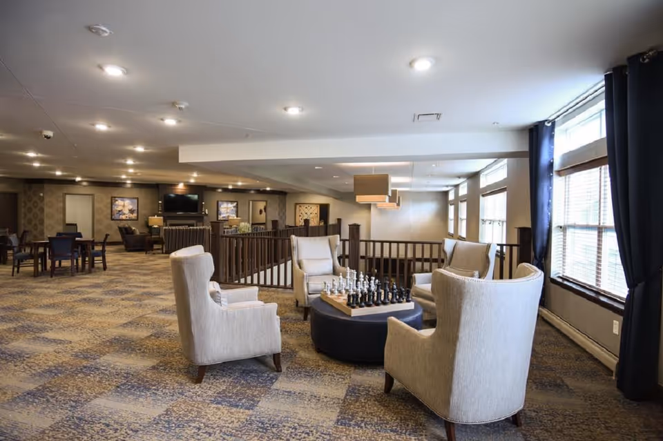 Open communal lounge with upholstered armchairs arranged around a chessboard table and windows along the right side.