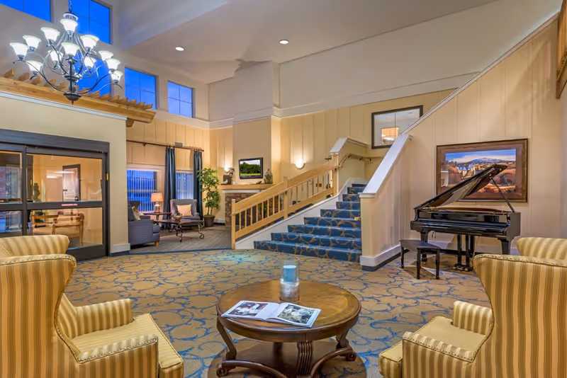 Spacious furnished lobby with seating areas, a grand piano, carpeted staircase and chandelier.