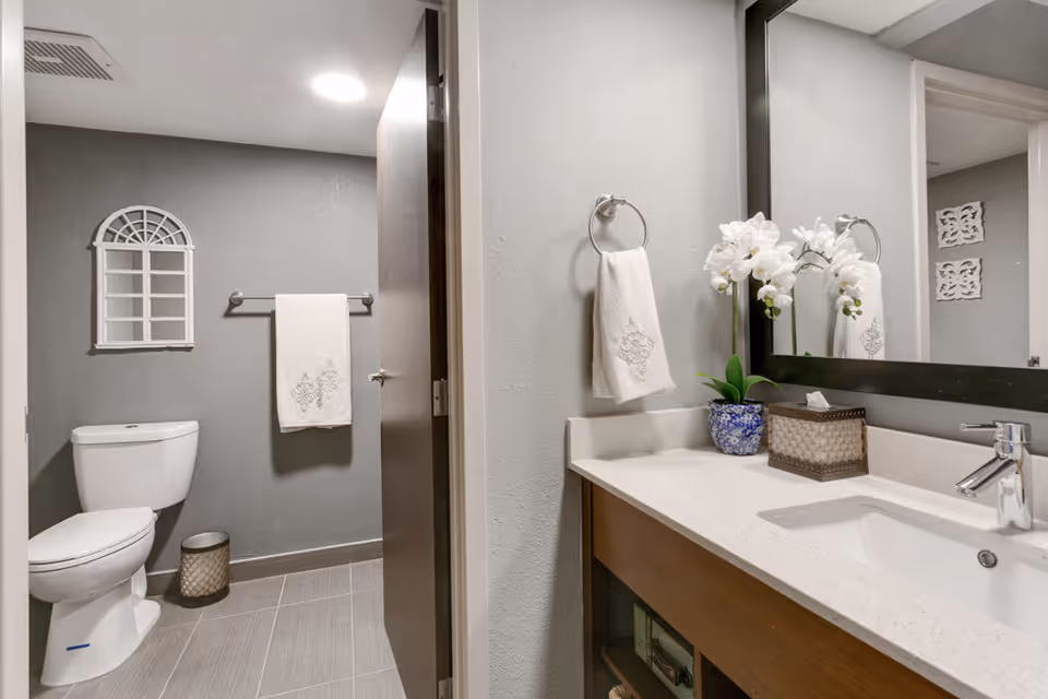 A modern bathroom featuring a white toilet, a towel hanging on a silver towel rack against a gray wall, and a vanity with a white countertop, a sink, a faucet, a hand towel on a ring, a blue and white flower pot with white orchids, and a tissue box. There is a large mirror above the vanity reflecting part of the room.