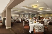 A spacious dining room with multiple tables covered in white tablecloths and set with plates, glasses, and napkins. The room has patterned upholstered chairs, carpeted floors, and ceiling lights. Large windows with curtains allow natural light to enter, and several people are seated and dining.