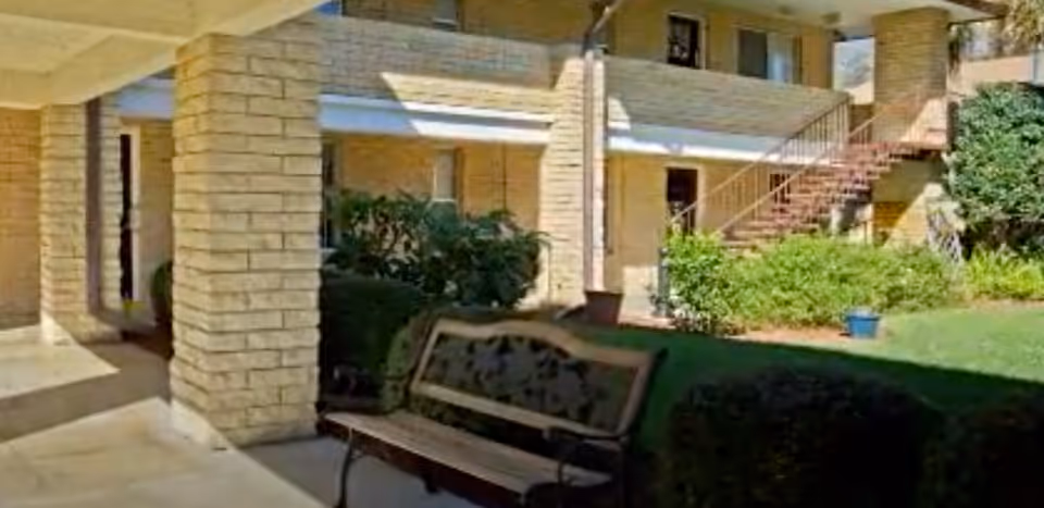 Outdoor courtyard area of a senior living facility with a bench, brick pillars, stairs leading to the second floor, and greenery including bushes and a potted plant.