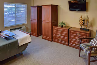 A furnished senior living bedroom with a bed, armoire, dressers, and a chair.