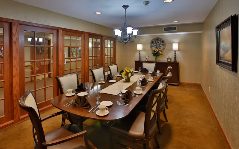 A formal dining room with a long wooden table set for eight people. The table has white cups and saucers, folded brown napkins, silverware, and glassware. There are two floral centerpieces on the table. The room features beige walls, a chandelier, two table lamps on a sideboard, and framed artwork on the walls. Wooden framed glass doors line one side of the room.