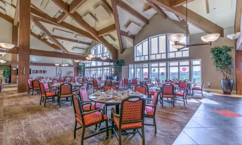 Spacious dining room with multiple tables and red cushioned chairs arranged neatly. The room features large arched windows allowing natural light to fill the space, wooden beams on a high ceiling, and several hanging light fixtures. There are plants in large pots placed near the windows and walls, and the floor is a combination of carpet and tile.