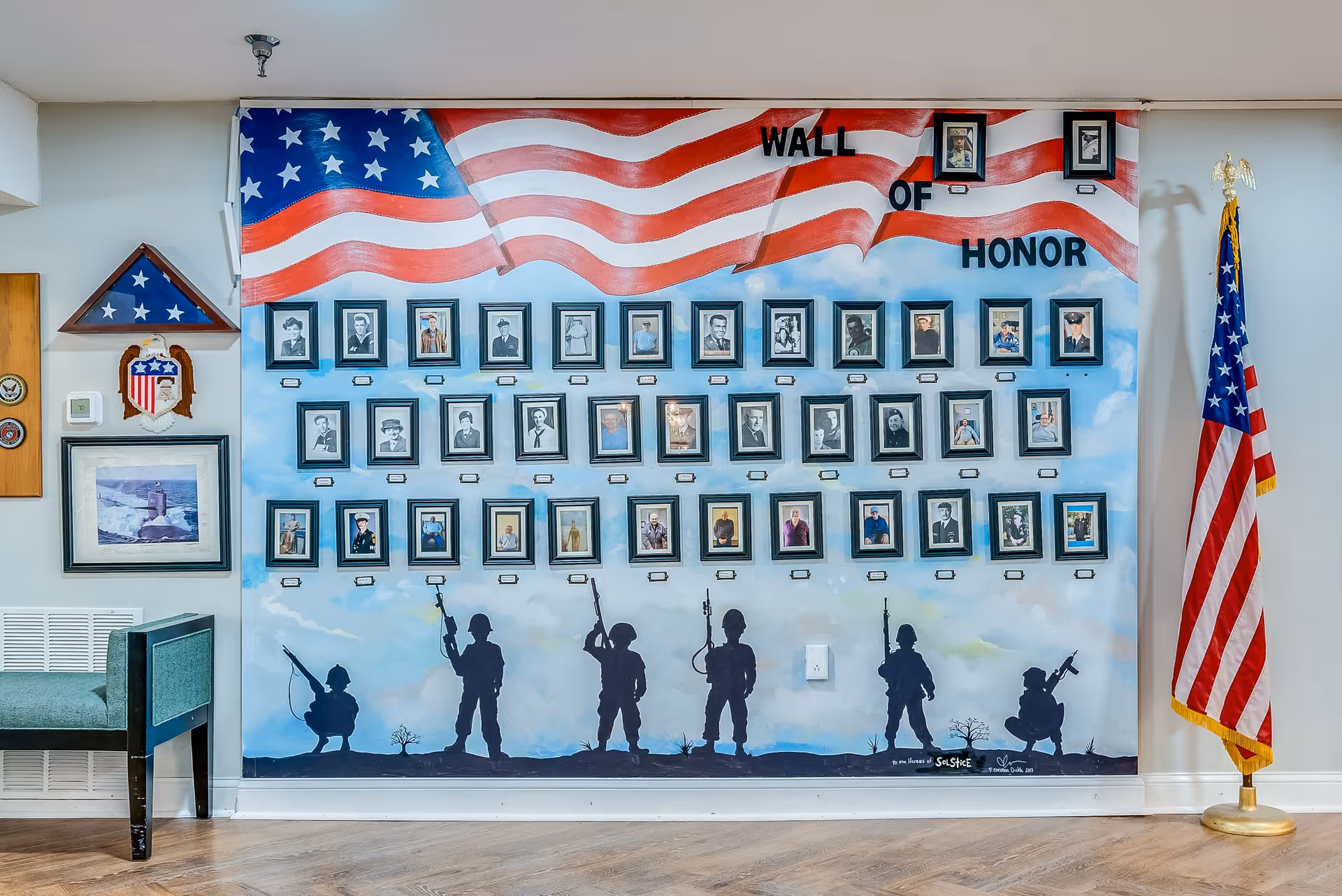 A Wall of Honor display featuring framed photographs of veterans arranged under a large painted American flag. Silhouettes of soldiers holding rifles are painted below the photos against a sky background. To the right of the display is a standing American flag, and to the left is a folded flag in a triangular case along with military insignia and a framed picture of a lighthouse. The floor is wooden, and there is a green bench partially visible on the left.