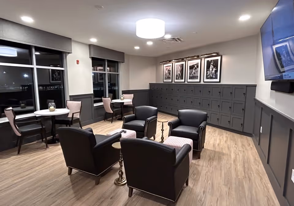 A cozy lounge area with four black armchairs arranged around two small round tables. Behind the chairs, there is a wall with multiple small lockers and four framed black-and-white photographs above them. To the left, there are two small round tables each with two chairs near large windows. The room has wood flooring, gray wainscoting on the walls, and a ceiling light fixture. A flat-screen TV is mounted on the right wall.