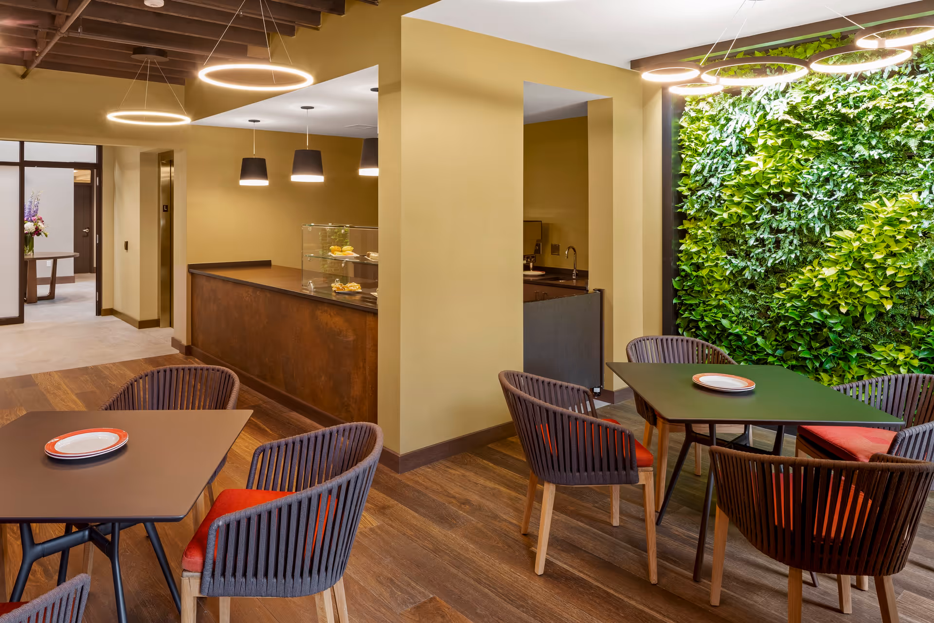 A modern dining area with two square tables, each surrounded by four chairs with red cushions. The room features a green living wall with plants on one side and warm yellow walls. Circular pendant lights hang from the ceiling, and there is a counter area with a glass display case containing pastries. The floor is wooden, and the ceiling has exposed wooden beams.