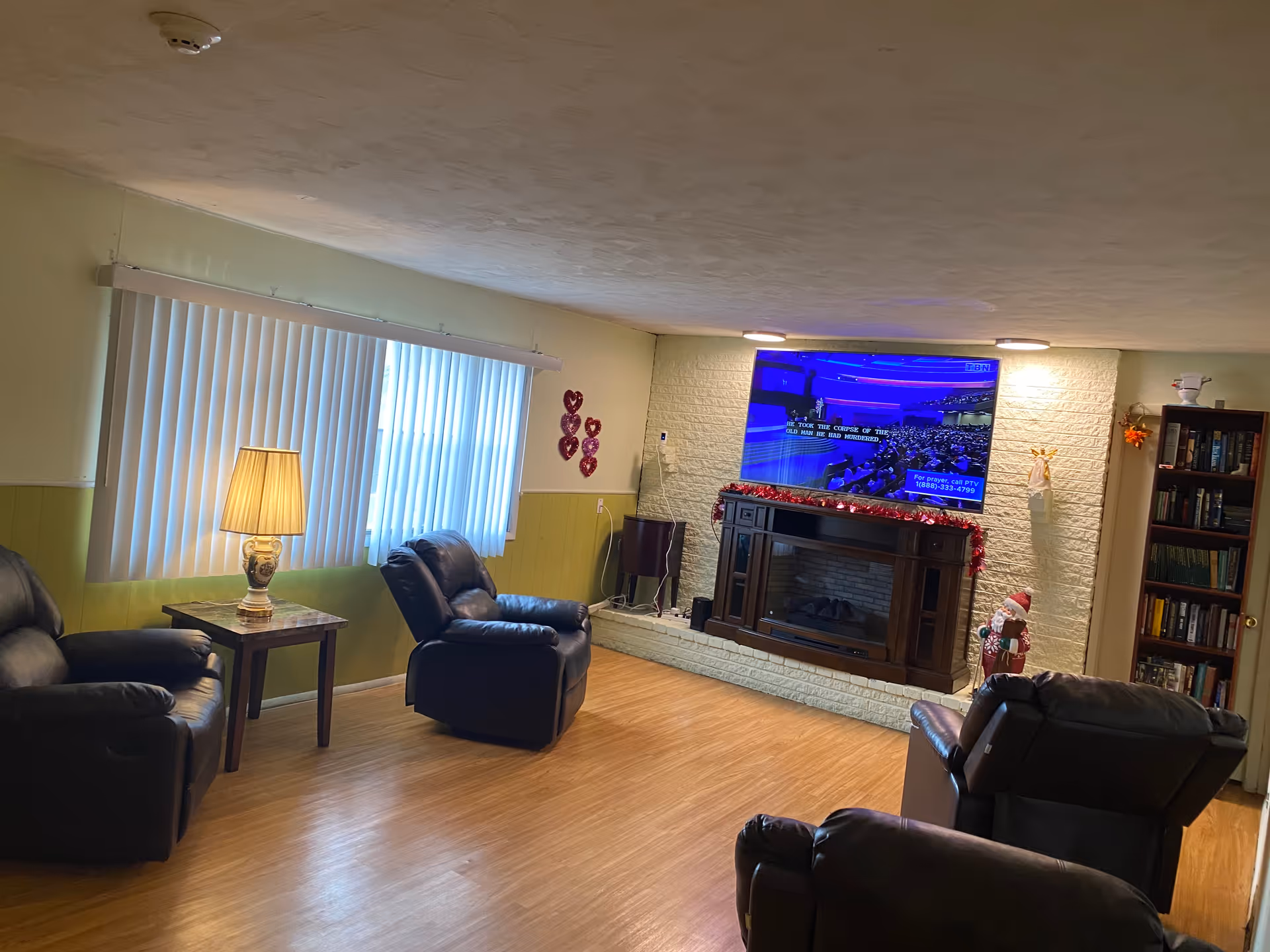 A cozy living room with four dark leather recliners arranged around a fireplace with a mounted flat-screen TV above it. The room has light green walls with a window covered by vertical blinds, a side table with a lamp, a bookshelf filled with books, and some holiday decorations including a Santa figure and red heart ornaments on the wall.
