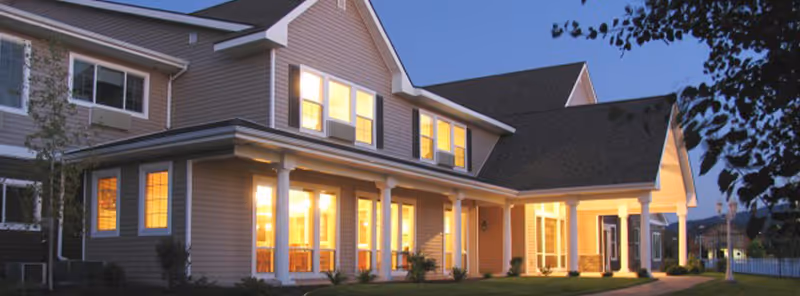 Exterior view of a two-story residential building at dusk with warm lights glowing from the windows and a covered porch supported by white columns. The building is surrounded by some landscaping and trees.