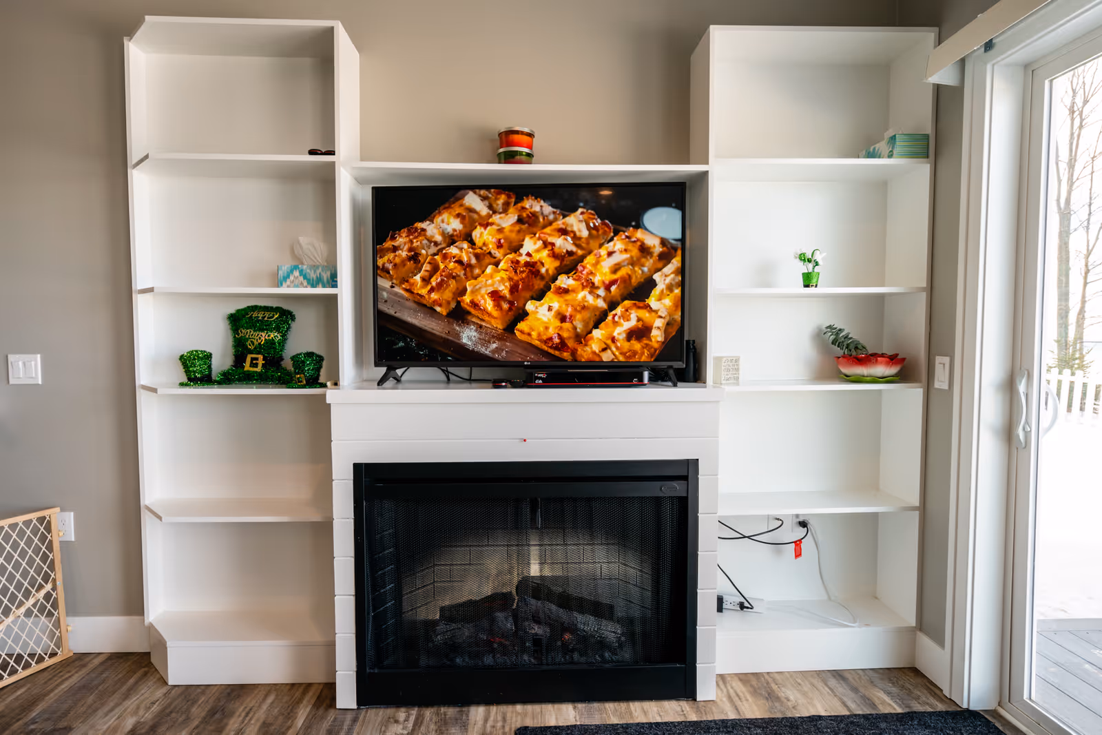 A cozy living room area featuring a white fireplace with a black screen, flanked by white built-in shelves on both sides. A flat-screen TV is mounted above the fireplace displaying an image of baked bread or pastry. The shelves hold a few decorative items including a green St. Patrick's Day hat and cups, a small plant, and a bowl. To the right, there is a sliding glass door leading outside, letting in natural light.