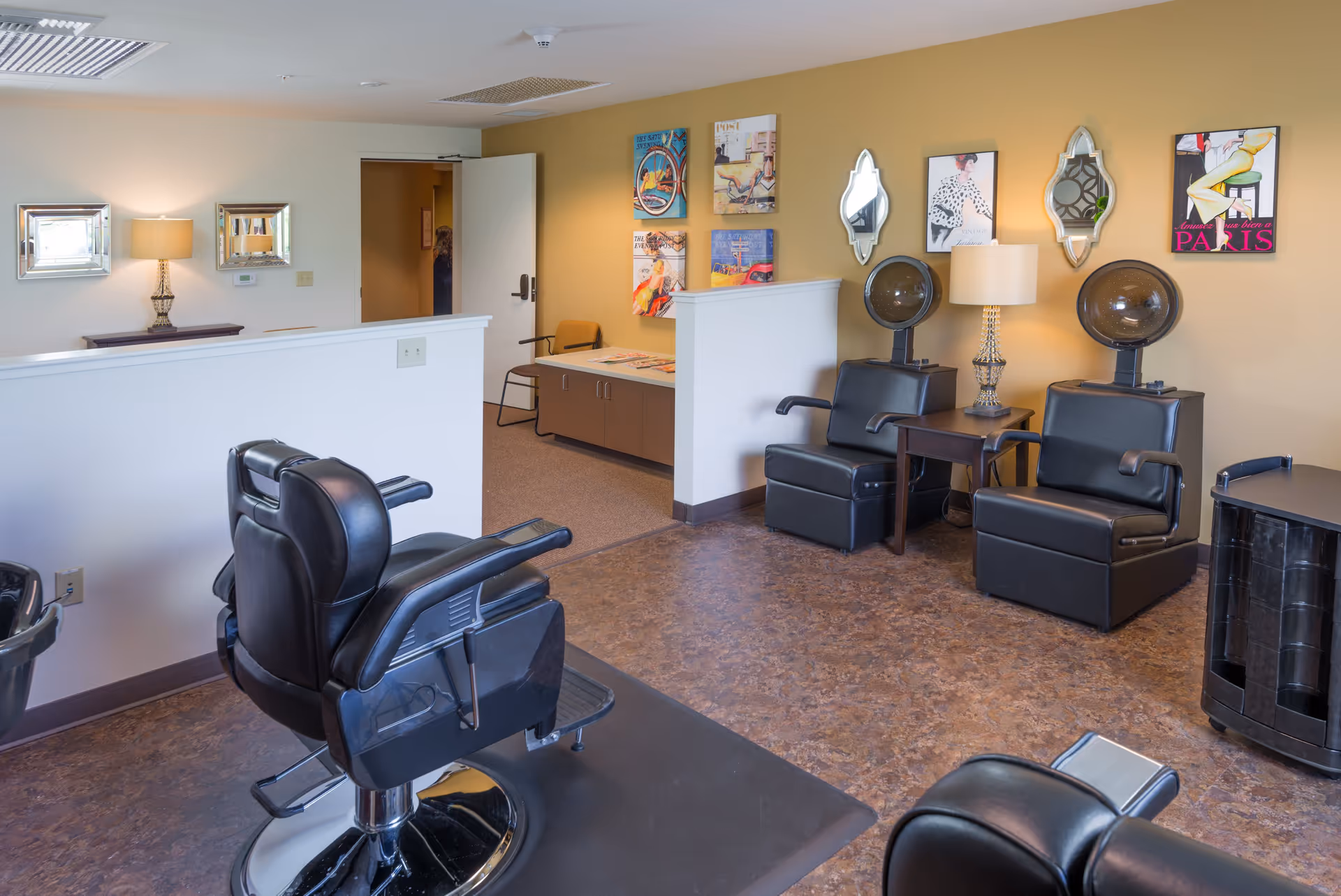 Interior of a senior living facility's salon area with black salon chairs, hair drying stations, a lamp on a side table, decorative mirrors, and colorful wall art. The room has a warm, inviting atmosphere with a mix of beige and white walls and a brown floor.