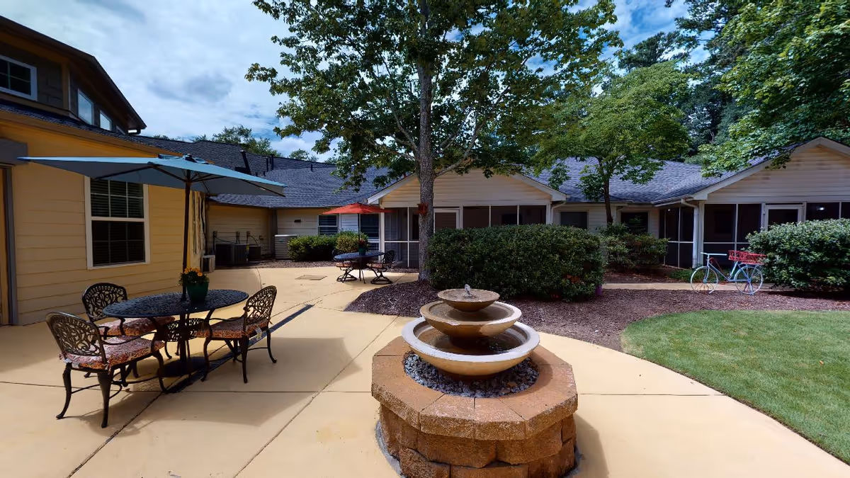 Outdoor patio area at Highlands Senior Living Norcross featuring a three-tiered stone water fountain in the center, surrounded by paved walkways. There are two round metal tables with chairs, each shaded by umbrellas, one blue and one red. The patio is bordered by bushes, trees, and a well-maintained lawn, with the building's exterior visible in the background under a partly cloudy sky.
