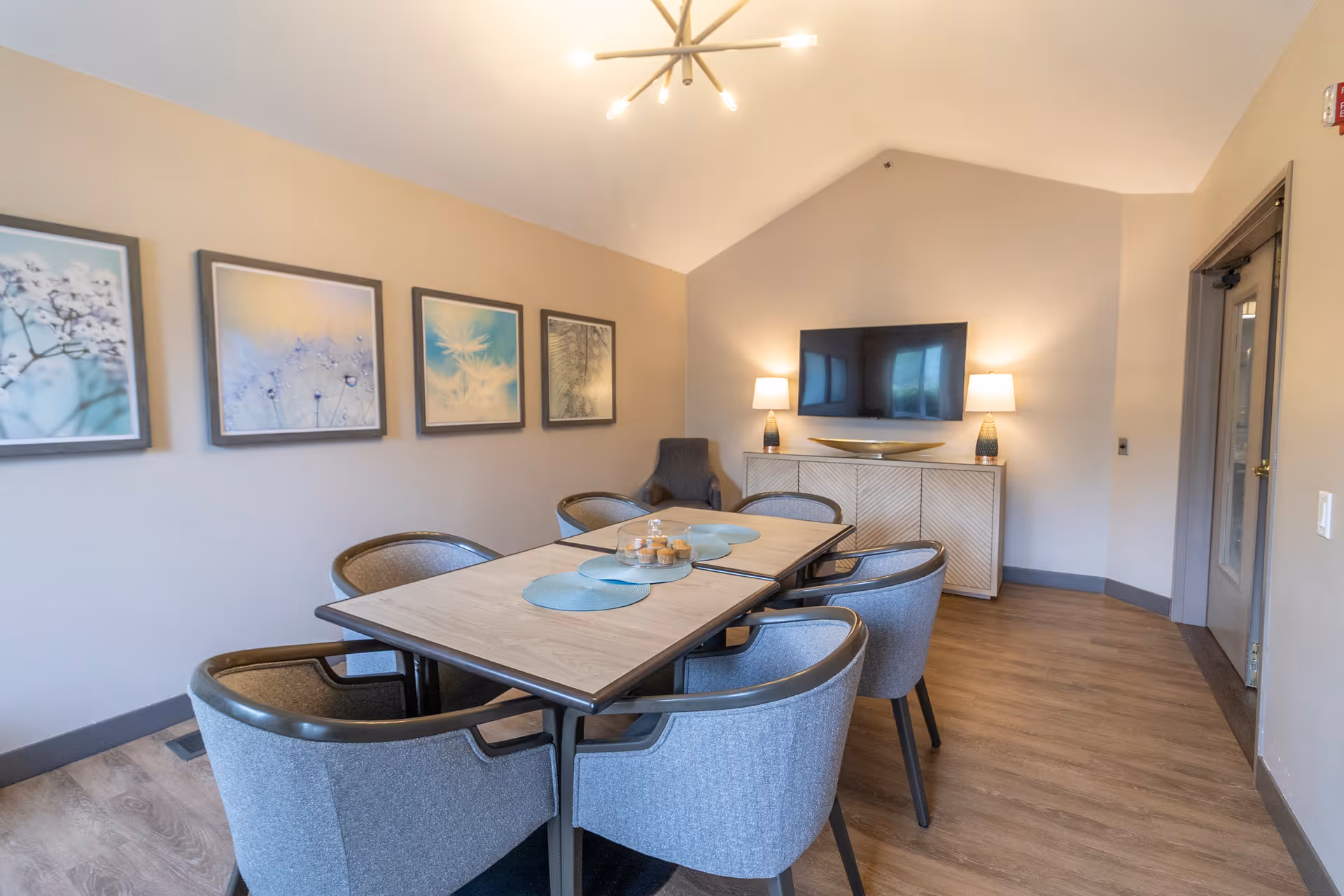 A modern dining room with a rectangular wooden table surrounded by six gray upholstered chairs. The table has blue placemats and a centerpiece with small jars. On the wall to the left, there are four framed pictures of nature scenes. At the far end of the room, there is a white sideboard with two table lamps and a large flat-screen TV mounted above it. The room has wood flooring and a door on the right side.