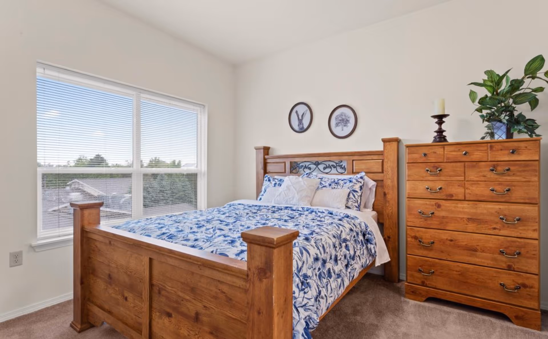 A bedroom with a wooden bed frame and matching wooden dresser. The bed is made with white and blue floral bedding and several pillows. Two framed pictures hang on the wall above the bed, and a green plant and candle are on top of the dresser. A large window with blinds lets in natural light.