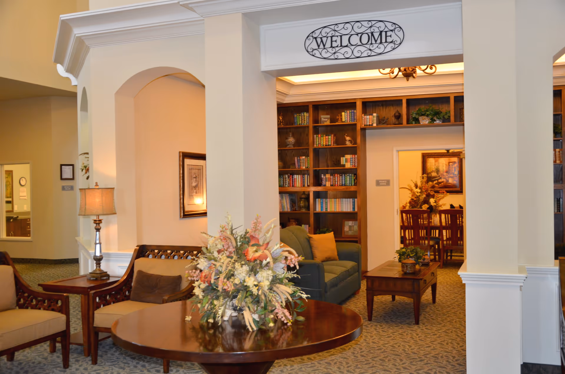 A cozy and warmly lit living room area with beige walls and carpeted floor. There are two cushioned armchairs with brown pillows, a round wooden table with a floral arrangement in the center, and a wooden coffee table with small plants. In the background, there is a green sofa with a yellow pillow, a large wooden bookshelf filled with books and decorative items, and a doorway leading to a dining room with a wooden dining table and chairs. Above the entrance to the living room area is a decorative sign that says 'WELCOME'.