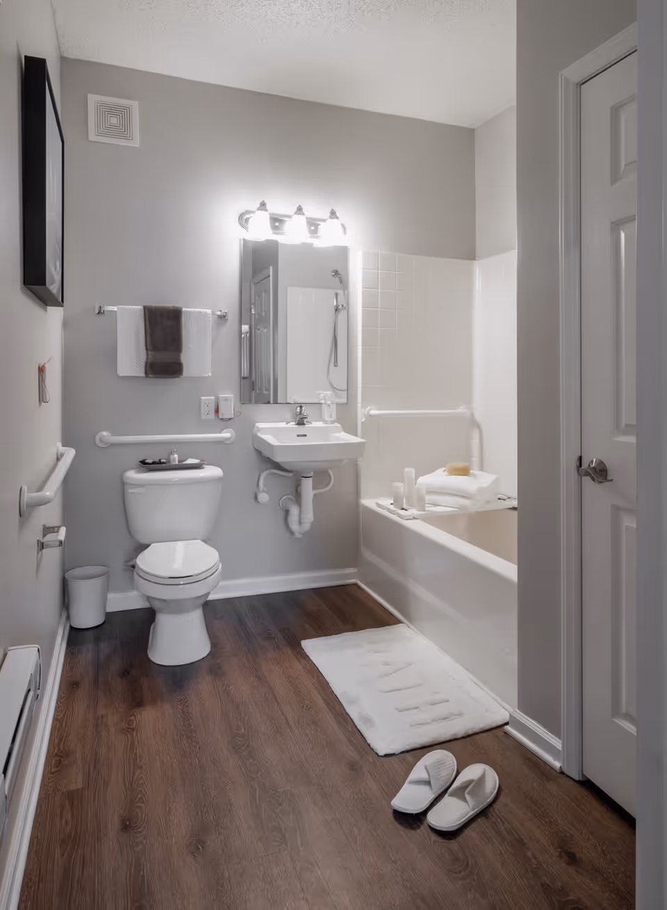 A clean and modern bathroom with a white toilet, a wall-mounted sink, a bathtub with white tiles, and grab bars for accessibility. There is a white bath mat on the wooden floor with a pair of white slippers next to it. A towel rack with two towels is mounted on the wall above the toilet, and a mirror with three lights is above the sink.