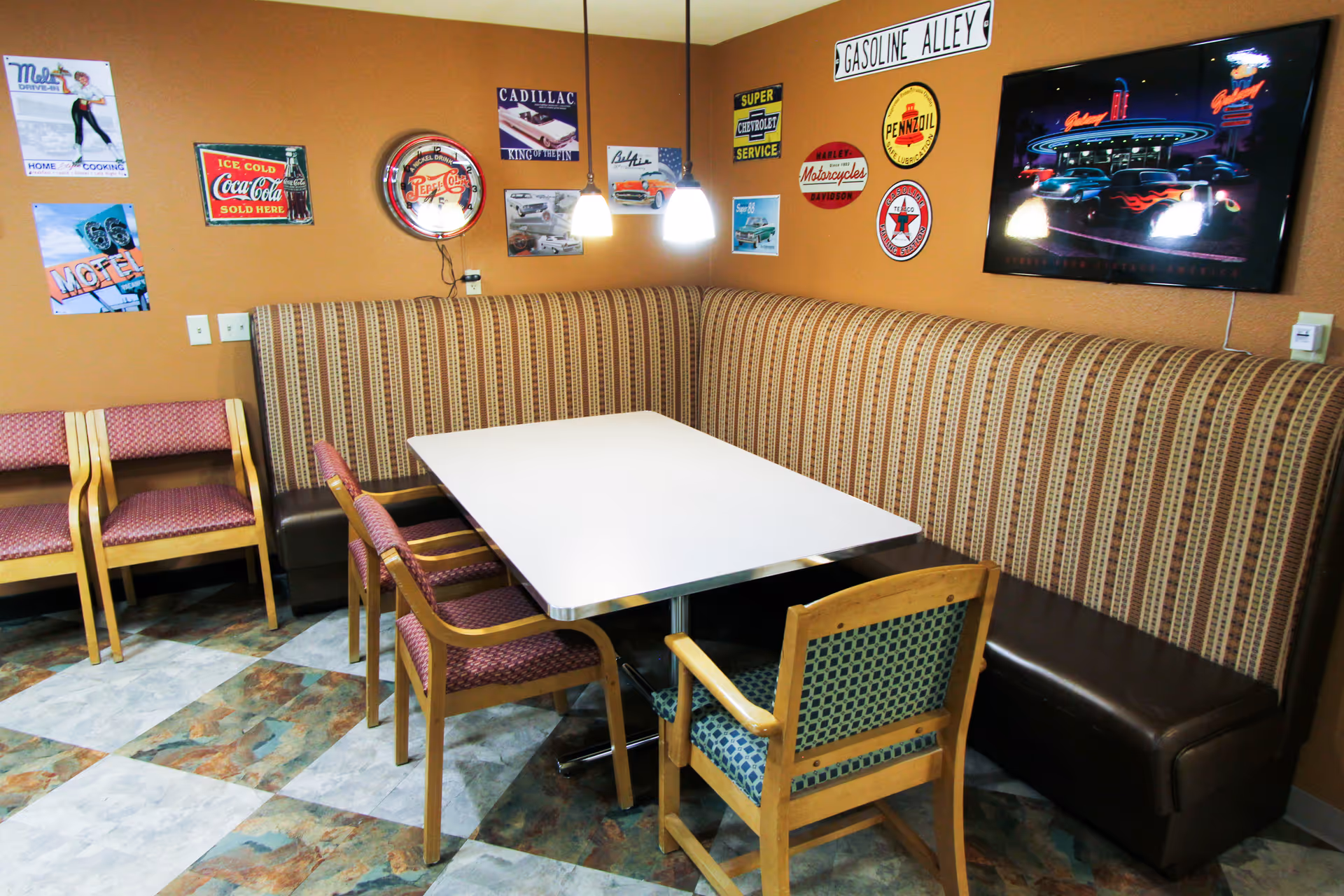 A cozy dining area with a white rectangular table surrounded by a striped upholstered bench and several wooden chairs with patterned cushions. The walls are decorated with vintage-style signs and posters, including advertisements for Coca-Cola, Pepsi-Cola, and various automotive brands. Two pendant lights hang above the table, and the floor has a patterned tile design.