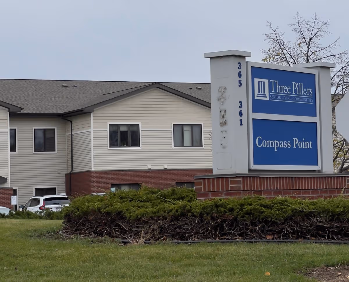 A Two-story senior living building with a lawn and shrubs and a prominent sign reading "Three Pillars Compass Point" in front.