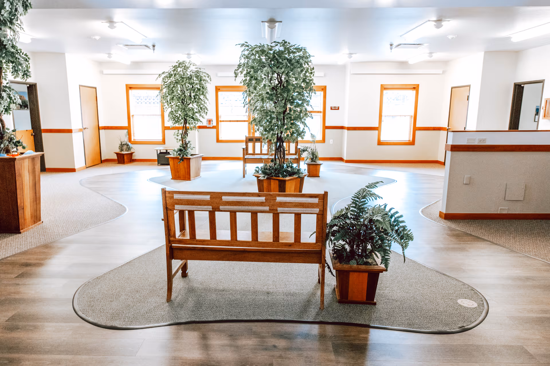 A bright and spacious interior common area with wooden benches and several potted plants. The room has large windows with natural light, light-colored walls with wooden trim, and a mix of carpet and wood flooring.