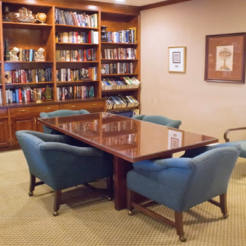 A cozy reading or meeting room with a polished wooden table surrounded by six blue upholstered chairs on casters. Behind the table is a large wooden bookshelf filled with books and decorative items, including a globe and small statues. The walls are beige with framed artwork hanging on them, and the floor is carpeted in a light pattern.