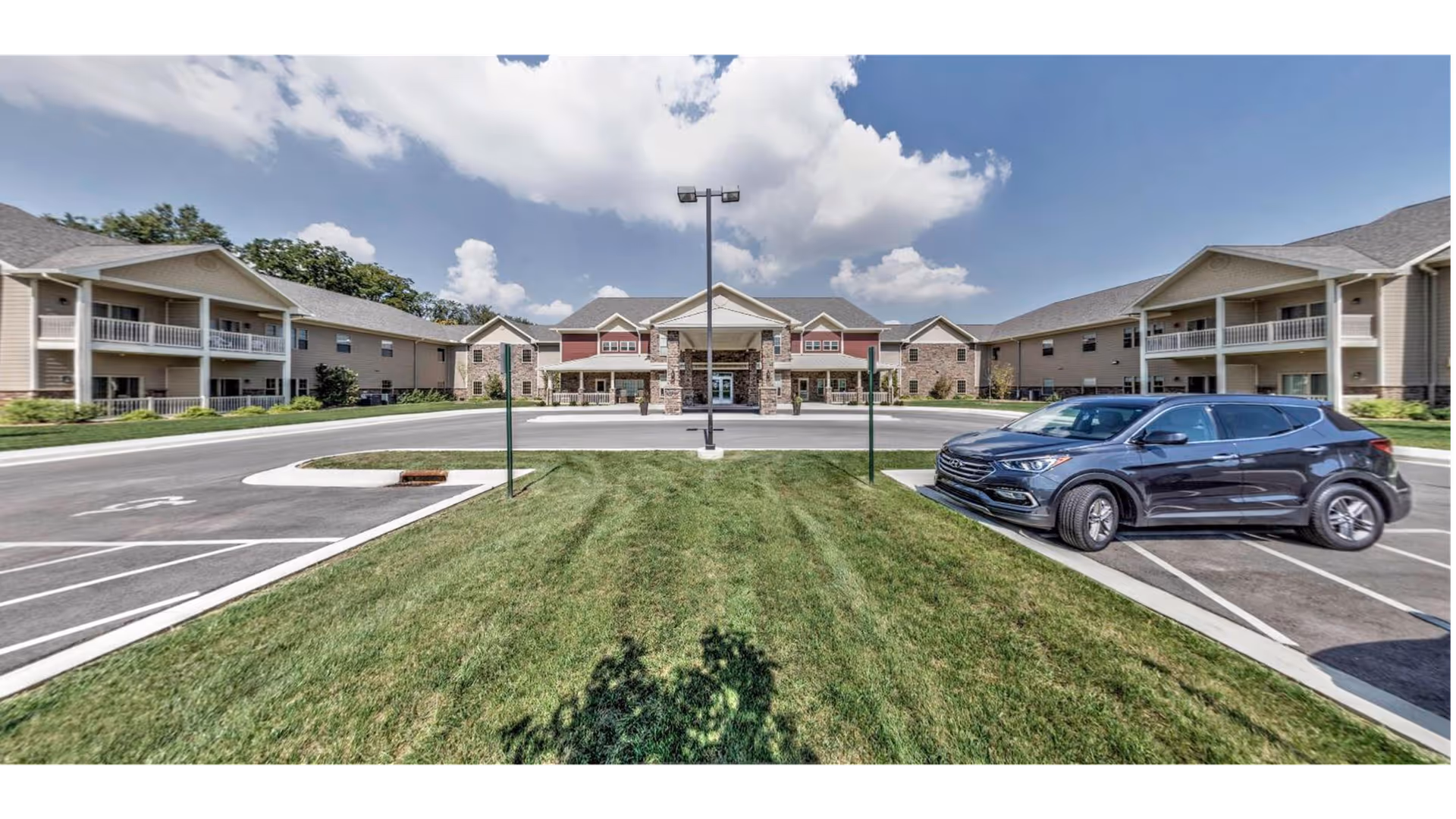 Front exterior view of Primrose Retirement Community of Appleton showing a large building with multiple sections, balconies, and a central entrance under a partly cloudy sky. There is a parking lot with a few cars and a grassy area in the foreground.
