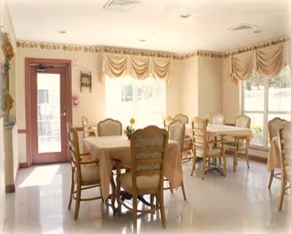 A bright dining room with several tables covered with beige tablecloths and surrounded by wooden chairs with cushioned seats. The room has large windows with beige valance curtains allowing natural light to fill the space. There is a door with a window on the left side and light-colored walls with a decorative border near the ceiling.