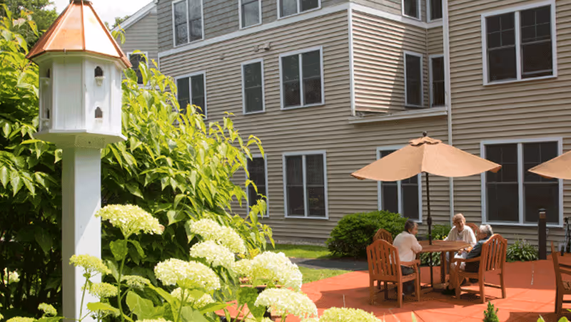 Three elderly people sitting and talking at a round wooden table with umbrellas on a patio outside a beige multi-story building, surrounded by green bushes and white flowers.