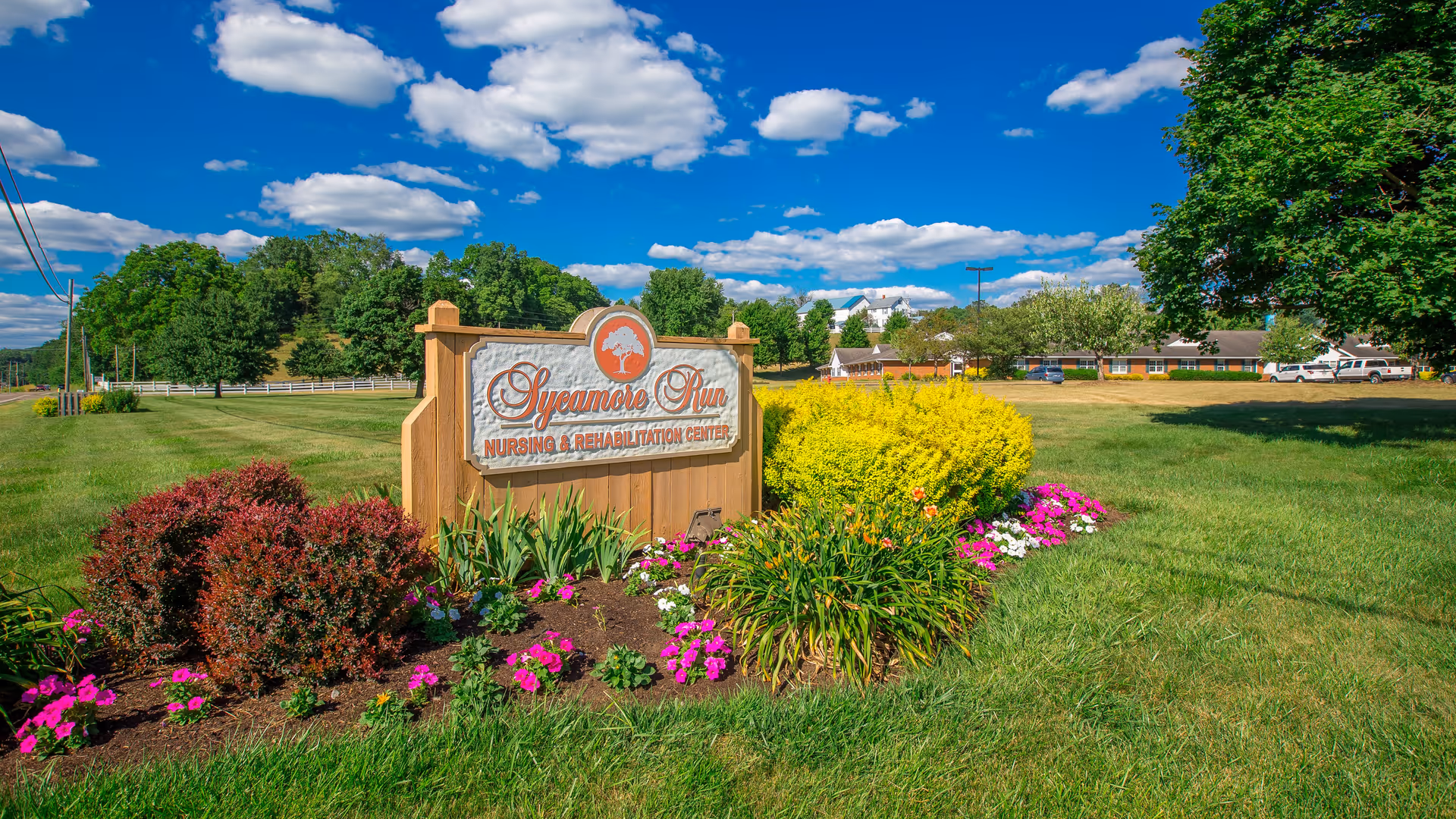 A wooden sign for Sycamore Run Nursing & Rehabilitation Center surrounded by colorful flowers and bushes on a grassy lawn under a blue sky with scattered clouds. Trees and part of the facility building are visible in the background.