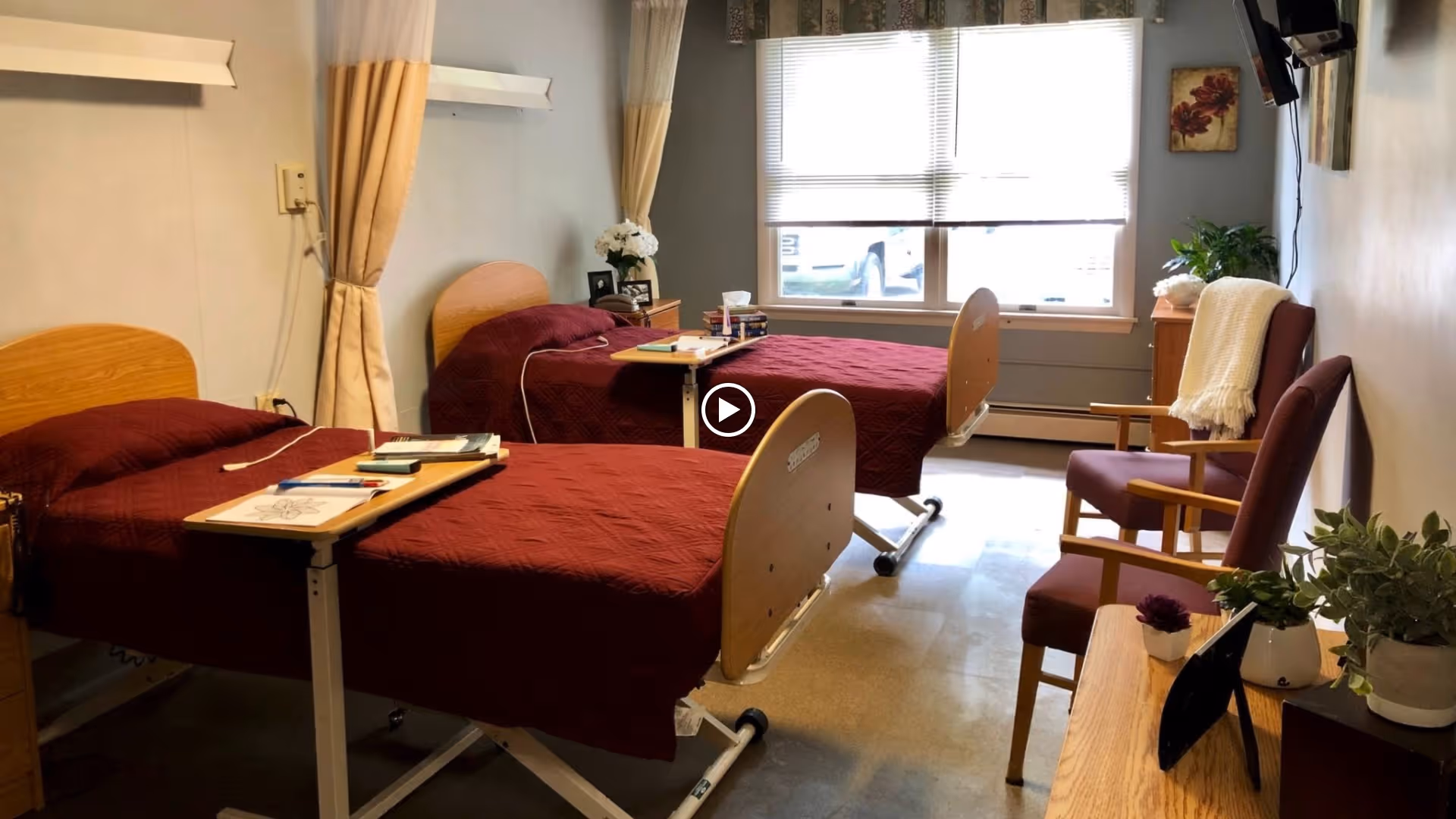 A senior living facility bedroom with two hospital-style beds covered in maroon quilts. Each bed has an overbed table with books and papers on it. There are two purple armchairs with wooden arms, a window with blinds letting in natural light, and several potted plants and decorations around the room.