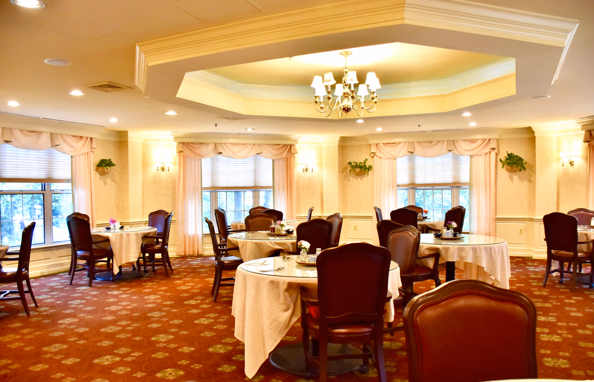 A well-lit dining room with round tables covered in white tablecloths, each surrounded by dark wooden chairs with cushioned seats. The room features large windows with pink curtains and valances, a patterned carpet, and a chandelier hanging from a recessed ceiling.