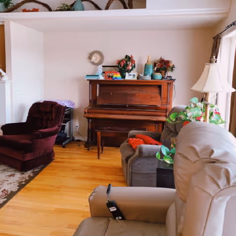 A cozy living room with a wooden piano against the wall decorated with various vases and flowers. There is a burgundy armchair on the left, a gray sofa with red cushions in the middle, and a beige recliner chair in the foreground. A floor lamp and some green plants are near a window on the right side.