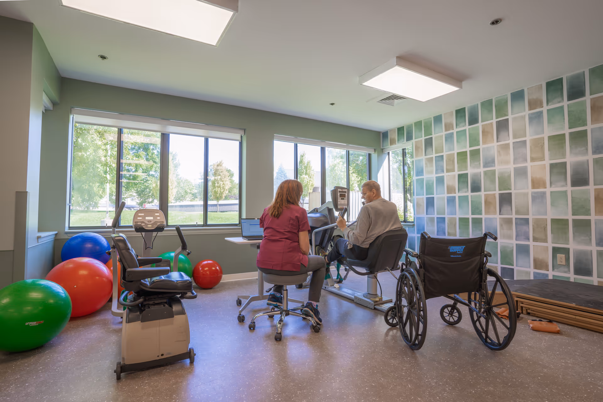 A physical therapy room with large windows showing greenery outside. A woman in a maroon uniform is sitting on a rolling stool next to an elderly man who is using a stationary exercise bike. There are several large exercise balls in red, green, and blue colors on the floor. A wheelchair is positioned nearby, and the wall has a decorative pattern of green and beige squares.