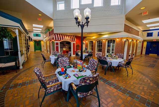 Indoor common area designed to look like a small town street with brick flooring, storefront facades, and tables with floral cushioned chairs set up for activities or dining. A vintage-style street lamp is in the center, and there are exit signs visible on the walls.