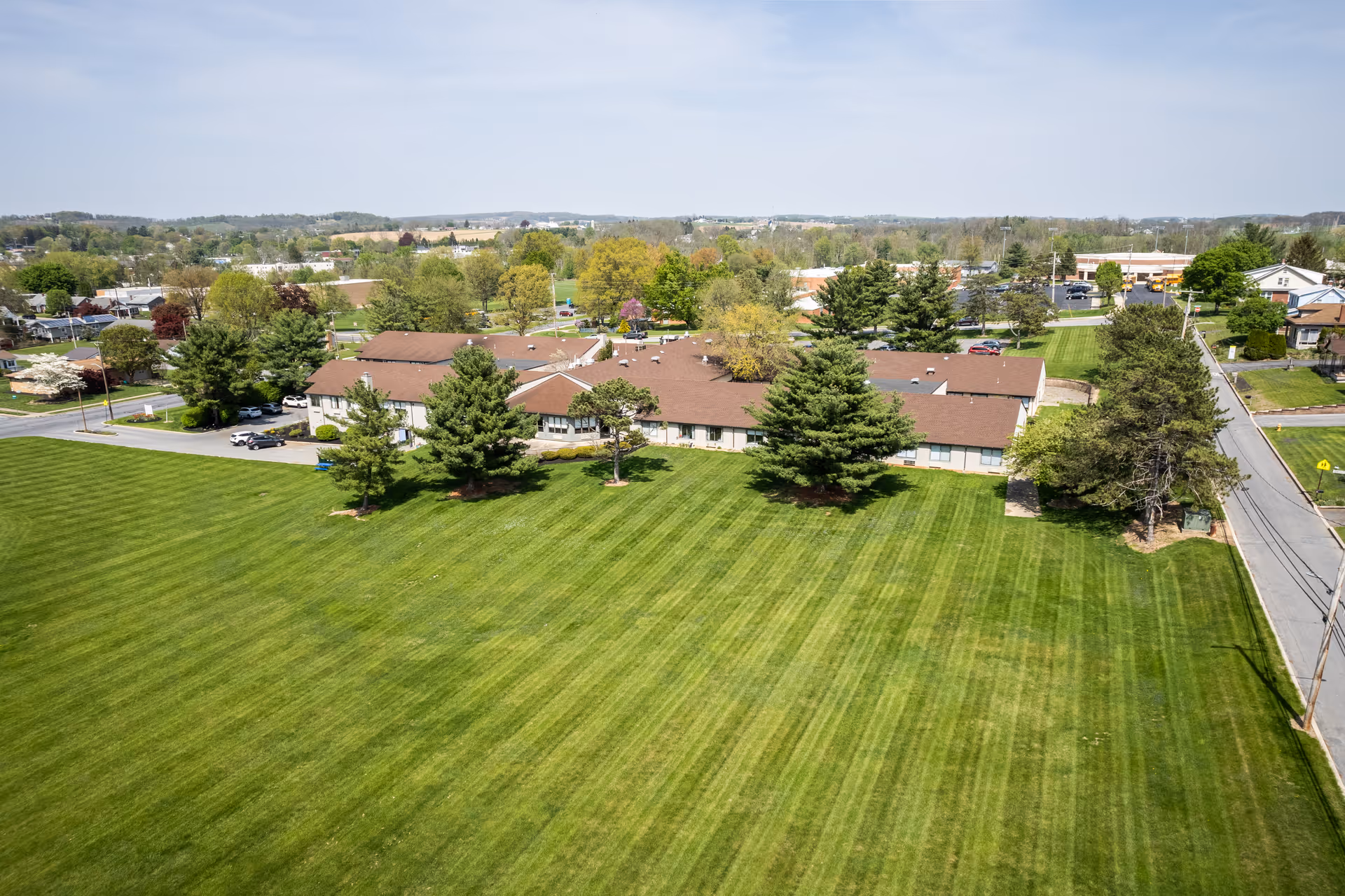 Aerial view of Willow Brook Rehabilitation and Healthcare Center showing a single-story building with a brown roof surrounded by a large, well-maintained green lawn with several trees. The surrounding area includes roads, houses, and other buildings under a clear sky.