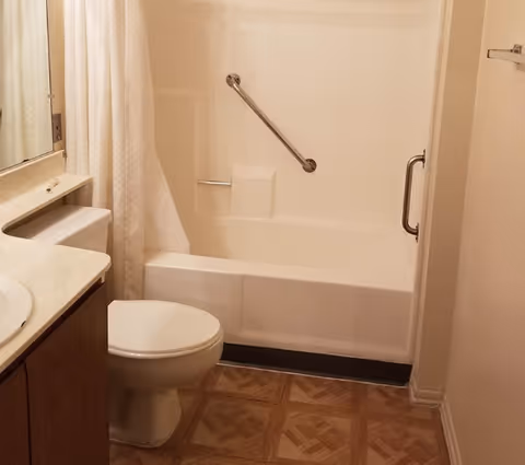 A small bathroom featuring a white bathtub with a grab bar and a shower curtain, a white toilet, a sink with a countertop, and a mirror above the sink. The floor has a patterned tile design.