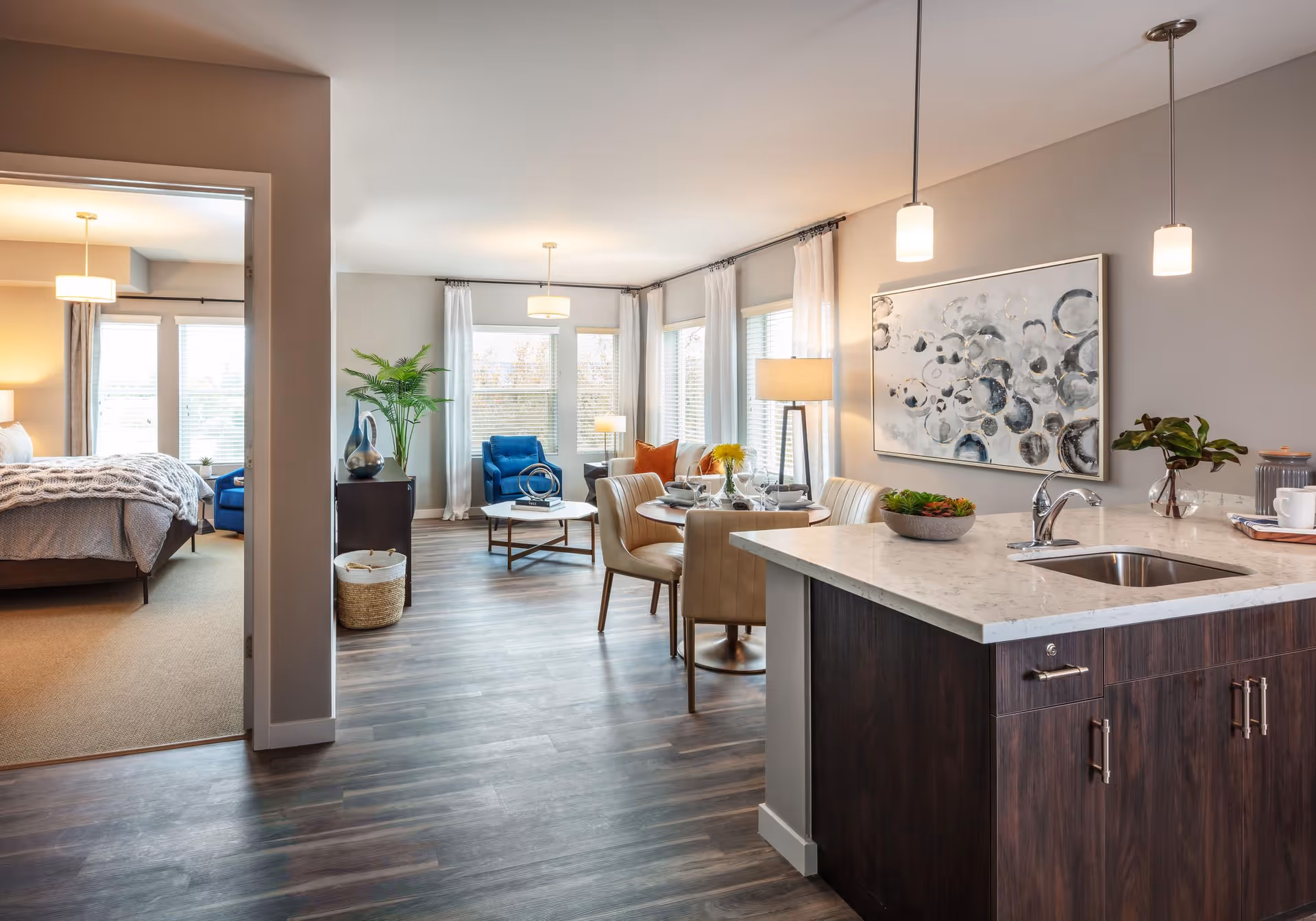 Open-concept apartment interior with a kitchen island in the foreground, a dining table and living area with chairs, and a bedroom visible through an open doorway.