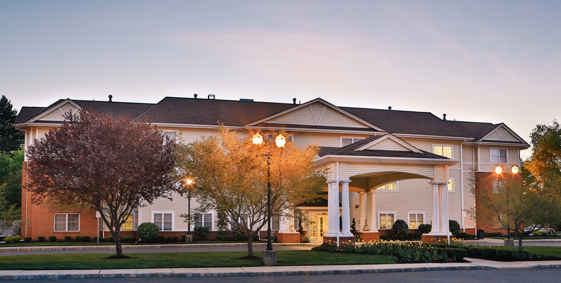 Exterior view of a senior living facility building named Brandywine Pennington by Monarch during dusk. The building has a beige facade with white trim and a covered entrance supported by white columns. There are trees with autumn-colored leaves and street lamps lit along the driveway in front of the building.