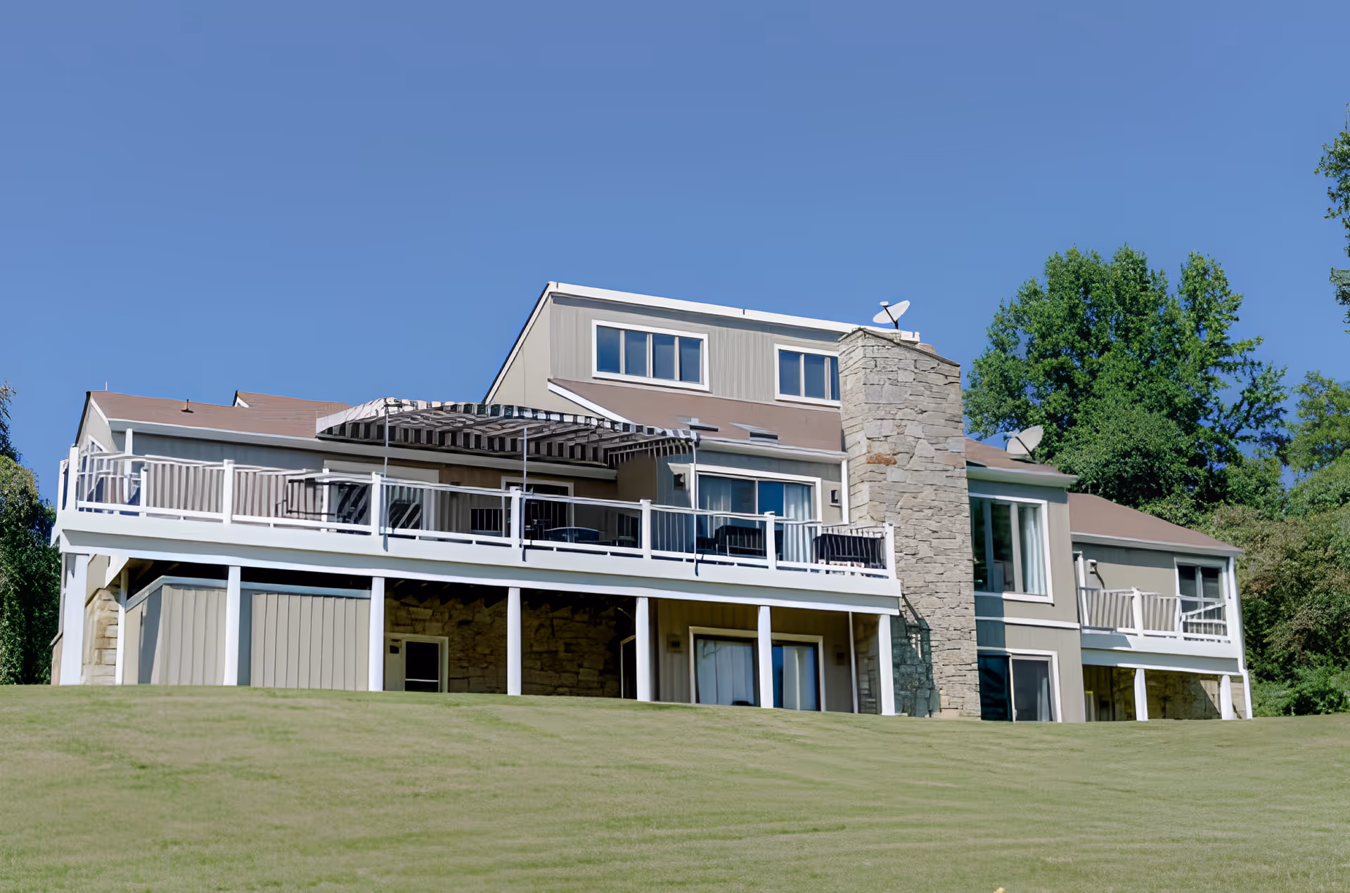 Exterior view of a multi-level assisted living facility building with beige siding, a stone chimney, multiple windows, and a large balcony with outdoor furniture and an awning. The building is set against a clear blue sky and surrounded by green trees and a grassy lawn.